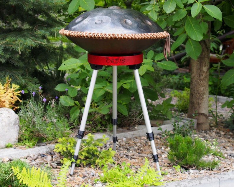 Puede incluir: Un handpan de acero con un borde tejido marr&oacute;n y una base roja, sostenido por un tr&iacute;pode blanco. La palabra "ORBIS" est&aacute; impresa en la base roja. El instrumento est&aacute; situado al aire libre, en medio de una exuberante vegetaci&oacute;n y un suelo cubierto de piedras.