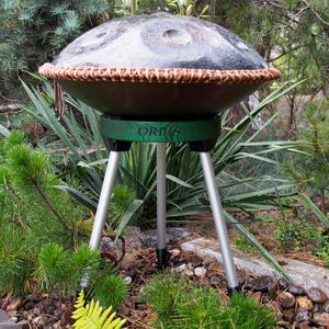 Puede incluir: Un handpan de metal oscuro con un ribete marr&oacute;n tejido, colocado sobre un soporte verde con la palabra "ORBIS" y tres patas plateadas. El instrumento est&aacute; al aire libre, rodeado de plantas verdes y rocas.