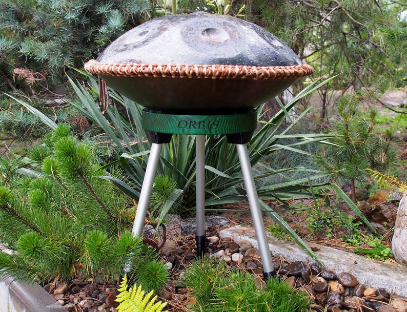 Puede incluir: Un handpan de metal oscuro con un ribete marr&oacute;n tejido, colocado sobre un soporte verde con la palabra "ORBIS" y tres patas plateadas. El instrumento est&aacute; al aire libre, rodeado de plantas verdes y rocas.