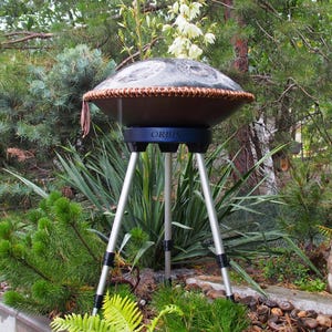 Puede incluir: Un handpan marr&oacute;n oscuro con un borde tejido, sostenido por un tr&iacute;pode. El soporte es negro y plateado, con la palabra "ORBIS" visible. El instrumento est&aacute; situado al aire libre en medio de una exuberante vegetaci&oacute;n.