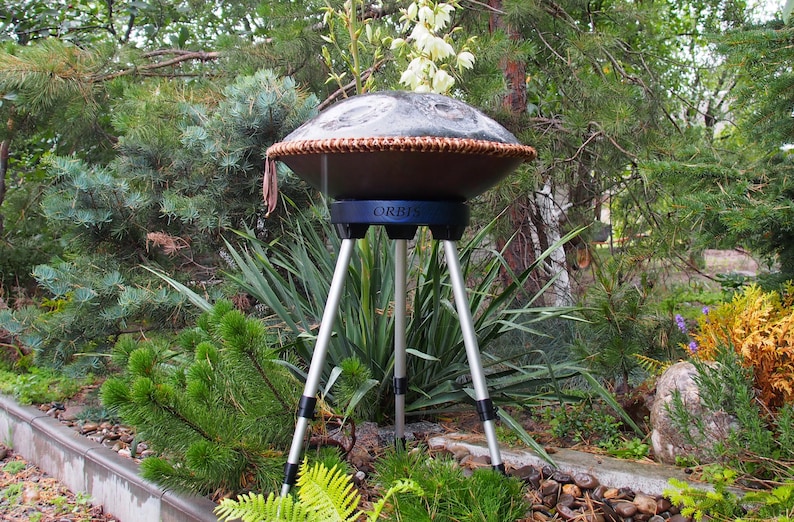 Puede incluir: Un handpan marr&oacute;n oscuro con un borde tejido, sostenido por un tr&iacute;pode. El soporte es negro y plateado, con la palabra "ORBIS" visible. El instrumento est&aacute; situado al aire libre en medio de una exuberante vegetaci&oacute;n.