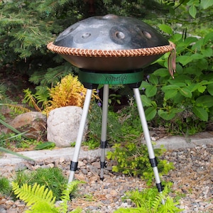 Puede incluir: Un handpan de acero con un borde tejido marr&oacute;n, apoyado en un soporte verde con la palabra "ORBIS" impresa. El soporte tiene tres patas plateadas. El instrumento est&aacute; al aire libre, rodeado de vegetaci&oacute;n y un camino de piedra.