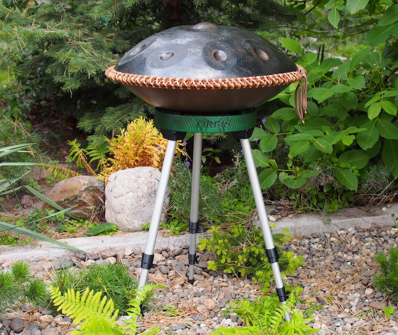 Puede incluir: Un handpan de acero con un borde tejido marr&oacute;n, apoyado en un soporte verde con la palabra "ORBIS" impresa. El soporte tiene tres patas plateadas. El instrumento est&aacute; al aire libre, rodeado de vegetaci&oacute;n y un camino de piedra.