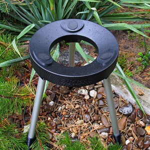 Puede incluir: Un soporte para plantas negro y circular con tres patas plateadas. El soporte tiene una abertura central y tres almohadillas circulares elevadas. El soporte est&aacute; colocado en un parterre con plantas verdes y peque&ntilde;as rocas.