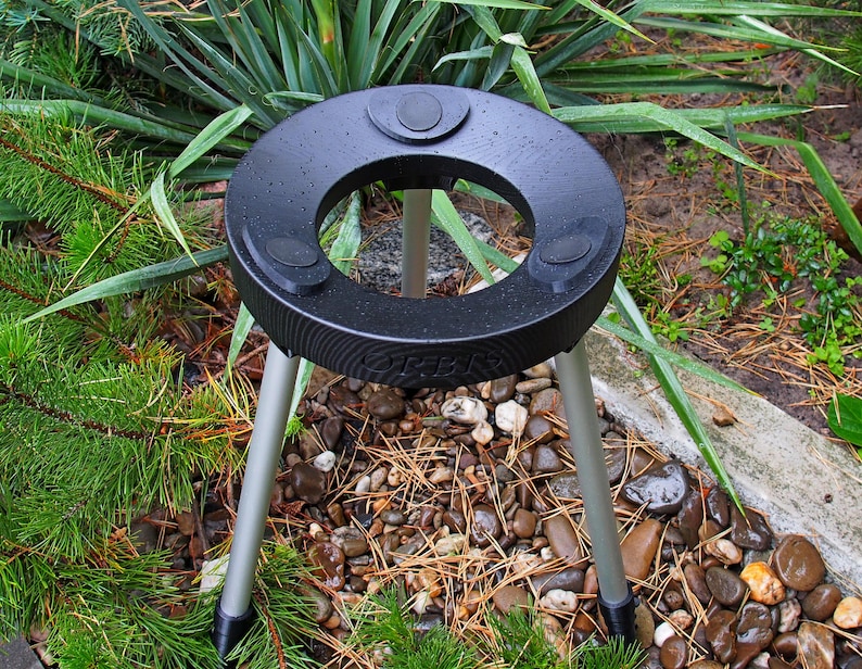 Puede incluir: Un soporte para plantas negro y circular con tres patas plateadas. El soporte tiene una abertura central y tres almohadillas circulares elevadas. El soporte est&aacute; colocado en un parterre con plantas verdes y peque&ntilde;as rocas.