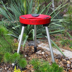 Puede incluir: Un tr&iacute;pode rojo y negro con la palabra "ORBIS" impresa en la parte superior. El tr&iacute;pode tiene tres patas plateadas y est&aacute; instalado al aire libre entre vegetaci&oacute;n y rocas.