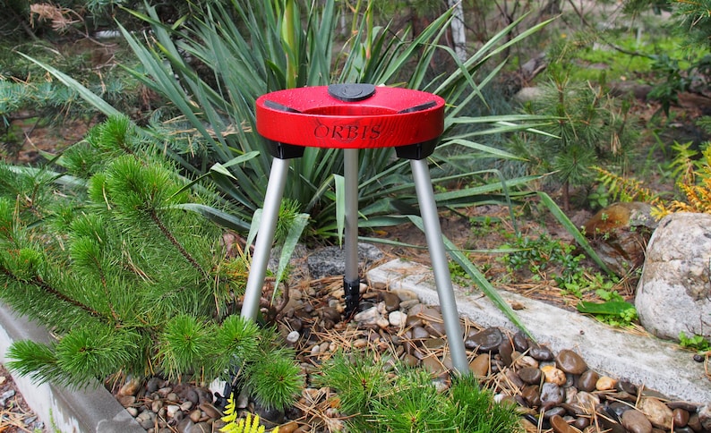 Puede incluir: Un tr&iacute;pode rojo y negro con la palabra "ORBIS" impresa en la parte superior. El tr&iacute;pode tiene tres patas plateadas y est&aacute; instalado al aire libre entre vegetaci&oacute;n y rocas.