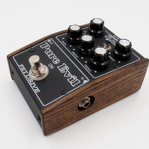 Puede incluir: Un pedal de efectos para guitarra Pure Evil FET Drive. El pedal tiene una carcasa de madera oscura con un panel superior negro. El panel presenta perillas para bajos, medios, agudos y drive, junto con un interruptor de pedal y conectores de entrada/salida.