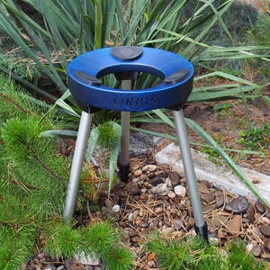 Puede incluir: Un tr&iacute;pode azul y negro con la palabra "ORBIS" impresa en el lateral. El soporte tiene tres patas plateadas y una parte superior circular con un centro negro. El soporte est&aacute; colocado en un jard&iacute;n con plantas verdes y peque&ntilde;as rocas.