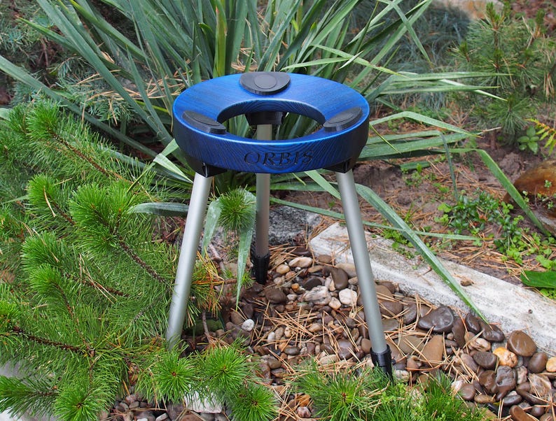 Puede incluir: Un tr&iacute;pode azul y negro con la palabra "ORBIS" impresa en el lateral. El soporte tiene tres patas plateadas y una parte superior circular con un centro negro. El soporte est&aacute; colocado en un jard&iacute;n con plantas verdes y peque&ntilde;as rocas.