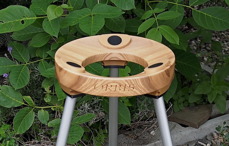 Puede incluir: Una mesa tr&iacute;pode de madera con una parte superior circular y tres patas. La parte superior tiene una abertura central y tres peque&ntilde;as hendiduras circulares. La palabra "ORBIS" est&aacute; grabada en la parte delantera. Las patas son plateadas.