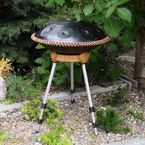Puede incluir: Un handpan de acero negro con un borde de cuero marr&oacute;n, apoyado sobre un soporte de madera con la palabra "VIRUS" impresa. El soporte est&aacute; sostenido por un tr&iacute;pode plateado. El instrumento est&aacute; al aire libre entre vegetaci&oacute;n.