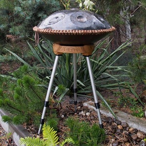 Puede incluir: Un handpan de acero gris oscuro con un borde tejido marr&oacute;n, montado sobre un soporte de madera con tres patas plateadas. El instrumento est&aacute; al aire libre, rodeado de vegetaci&oacute;n.