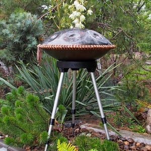 Puede incluir: Un handpan de acero marr&oacute;n oscuro con un borde tejido, colocado sobre un soporte negro con tres patas plateadas. El tambor est&aacute; rodeado de un exuberante follaje verde, incluyendo pinos y otras plantas.
