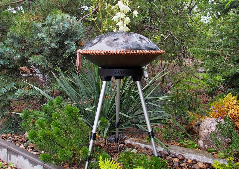 Puede incluir: Un handpan de acero marr&oacute;n oscuro con un borde tejido, colocado sobre un soporte negro con tres patas plateadas. El tambor est&aacute; rodeado de un exuberante follaje verde, incluyendo pinos y otras plantas.