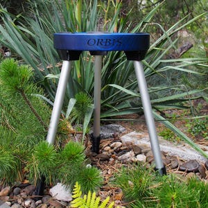 Puede incluir: Un tr&iacute;pode azul y negro con la palabra "ORBIS" impresa en la parte superior. El soporte tiene tres patas plateadas y est&aacute; situado en un jard&iacute;n con plantas verdes y peque&ntilde;as rocas.