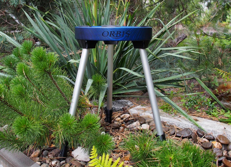 Puede incluir: Un tr&iacute;pode azul y negro con la palabra "ORBIS" impresa en la parte superior. El soporte tiene tres patas plateadas y est&aacute; situado en un jard&iacute;n con plantas verdes y peque&ntilde;as rocas.