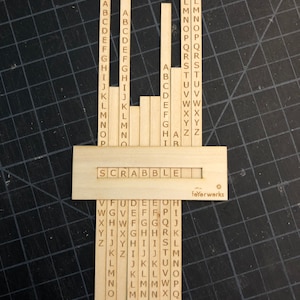 May include: Wooden Scrabble tiles with the letters of the alphabet printed on them. The tiles are arranged in columns, with each column representing a different letter. The tiles are on a wooden board with the word "SCRABBLE" printed on it.