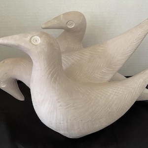 Op de afbeelding: Een sculptuur van drie gestileerde vogels in een lichtbeige kleur. De vogels hebben lange halzen en spitse snavels. De sculptuur is gemaakt van een textuurrijk materiaal en staat op een zwart oppervlak.