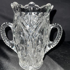 May include: Clear glass vase with two handles and a scalloped rim. The vase features an ornate, embossed pattern of floral and geometric designs. The glass is transparent, allowing light to pass through, and the vase sits on a round base.