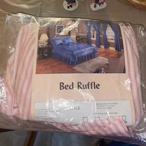May include: A packaged bed ruffle with a pink and white striped pattern. The packaging features an image of a blue bed set with matching curtains. The text "Bed Ruffle" is printed on the packaging.