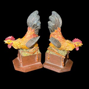 May include: A pair of rooster bookends. The roosters are yellow, orange, and red with gray tail feathers and red combs. They are perched on brown rectangular bases with a grassy texture.