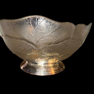May include: A clear glass bowl with a leaf design and a silver-colored base. The bowl has a scalloped rim and is likely used for serving or display. The leaf pattern adds a decorative touch to the bowl.