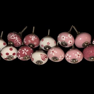 May include: A collection of sixteen round ceramic cabinet knobs featuring various pink and white patterns, including hearts, polka dots, and floral designs. Each knob has a metallic silver-toned base and a short threaded metal screw for mounting.