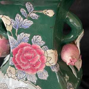 May include: A green ceramic vase with floral designs in pink, white, and blue. The vase has two handles and a square base.