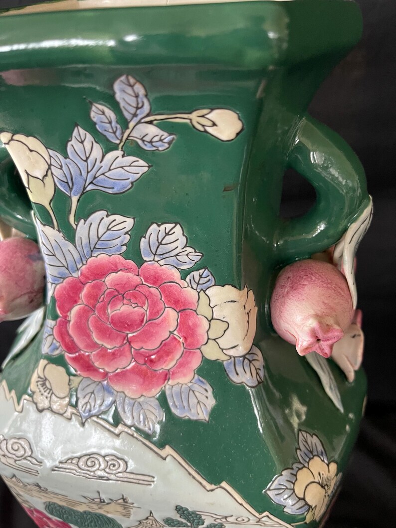 May include: A green ceramic vase with floral designs in pink, white, and blue. The vase has two handles and a square base.