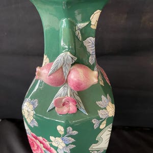 May include: A green ceramic vase with a hexagonal shape and floral designs. The vase has a raised design of pink fruit and leaves on the neck.