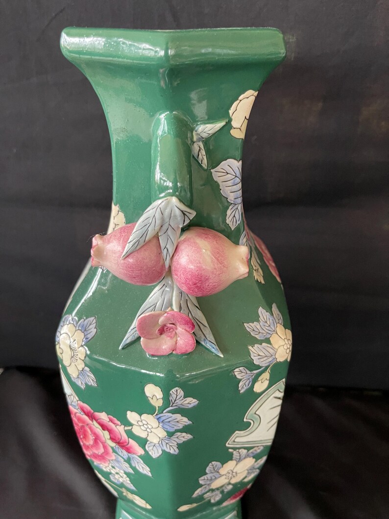 May include: A green ceramic vase with a hexagonal shape and floral designs. The vase has a raised design of pink fruit and leaves on the neck.