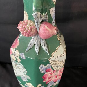 May include: A green ceramic vase with a floral design and a 3D pomegranate handle. The vase is decorated with pink, white, and blue flowers and leaves.