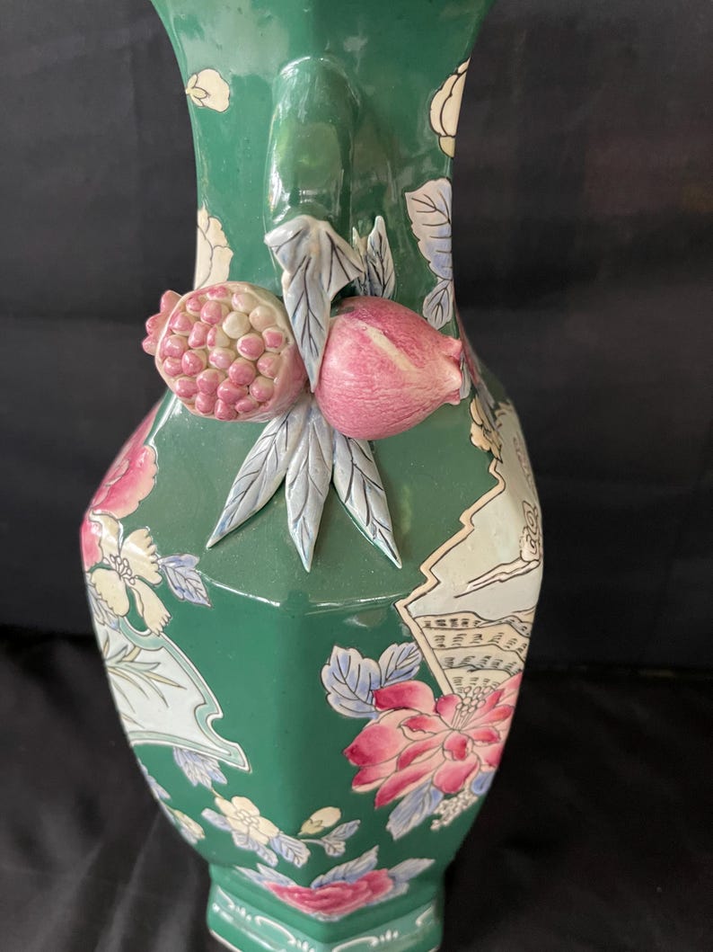 May include: A green ceramic vase with a floral design and a 3D pomegranate handle. The vase is decorated with pink, white, and blue flowers and leaves.