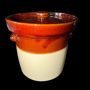May include: A two-tone ceramic crock with a lid. The upper portion is a glossy, reddish-brown, transitioning to a lighter tan color on the lower half. The lid has a small knob and the crock has two small handles.