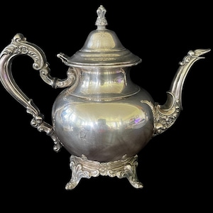 Puede incluir: Una tetera plateada con detalles ornamentados. La tetera presenta un asa curvada, un pico con adornos decorativos y una tapa rematada con un pequeño adorno. Se apoya sobre una base decorativa con tres patas, sobre un fondo negro.