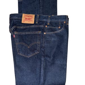 Könnte beinhalten: Dunkelblaue Jeans aus Denim mit einem beigen Lederaufnäher am hinteren Bund, der den Text Levi Strauss & Co. Quality Clothing trägt. Die Jeans hat kupferfarbene Nieten, orangefarbene Kontrastnähte und ein kleines rotes Etikett an der Tasche.