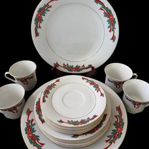 Puede incluir: Un juego de vajilla de porcelana blanca con un patrón festivo de cintas rojas, ramas de pino verdes y flores de pascua con bordes dorados. El conjunto incluye un plato grande, una pila de platillos pequeños y cuatro tazas a juego.