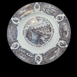 Peut inclure: Une assiette en céramique blanche avec une illustration bleue et marron d'une scène de rivière avec un bateau à vapeur et des bâtiments. L'assiette a une bordure décorative avec des vignettes historiques et le texte "1850 Town of Kansas, 1853 Centennial, 1880 City of Kansas."