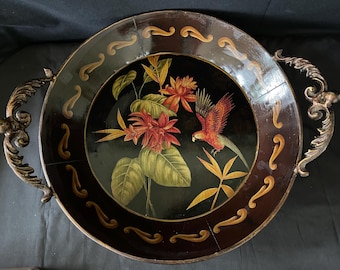 May include: A round decorative tray with a dark brown rim and a black interior. The tray features a painted design of a parrot, flowers, and foliage. The tray has ornate gold-colored handles on either side.