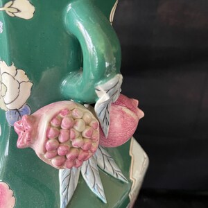 May include: A green ceramic vase with a floral design and a handle shaped like a pomegranate. The vase has a textured surface and is decorated with pink, white, and blue flowers.