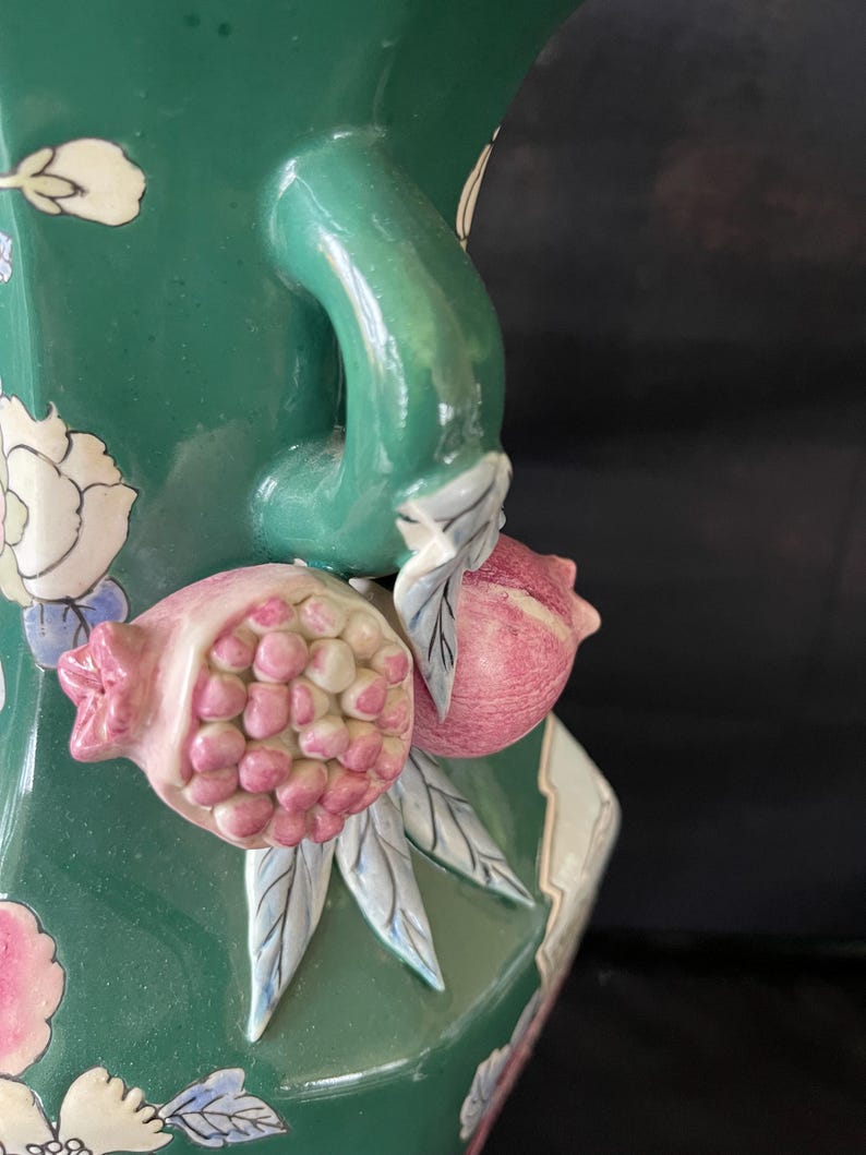 May include: A green ceramic vase with a floral design and a handle shaped like a pomegranate. The vase has a textured surface and is decorated with pink, white, and blue flowers.