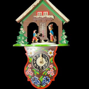 Puede incluir: Un reloj de pared estilo chalet con un termómetro en el centro, flanqueado por dos figuras sobre esquís. La sección inferior muestra una esfera con números arábigos, decorada con flores pintadas en blanco y azul sobre un fondo marrón.