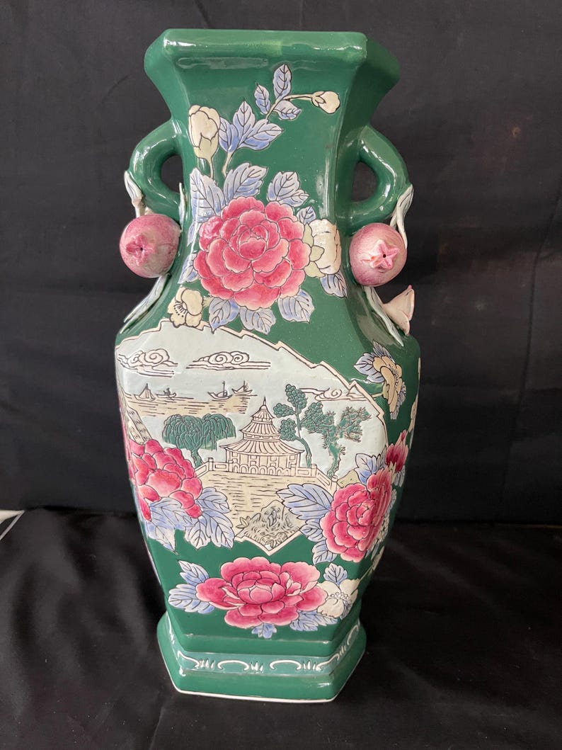 May include: A green ceramic vase with floral and landscape designs. The vase has two handles with pink fruit accents.