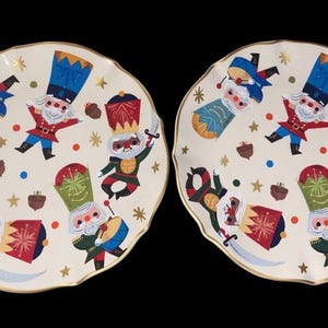 Puede incluir: Dos platos de cerámica blanquecina con borde festoneado y ribete dorado. Cada plato presenta un diseño caprichoso de soldados de juguete en varias poses, con detalles en azul, rojo, verde y dorado. Bellotas y estrellas completan el tema festivo navideño.