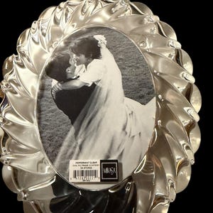May include: An oval clear glass picture frame with a swirling, textured border. A black and white photograph of a couple embracing is inside. A label reads: PEPPERMINT CLEAR OVAL PIC FRAME 10 3/4" 50801 SA 972/829 MIKASA, with a barcode.