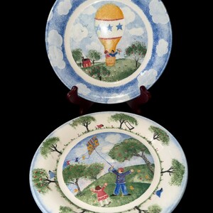 May include: Two round ceramic plates featuring hand-painted scenes. The top plate displays a yellow hot air balloon with blue stars over a green landscape. The bottom plate shows two figures flying a colorful kite in a park with trees and grass.