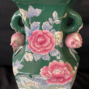 May include: A green ceramic vase with floral designs and two handles. The vase is decorated with pink and white flowers and green leaves. The handles are shaped like pomegranates.