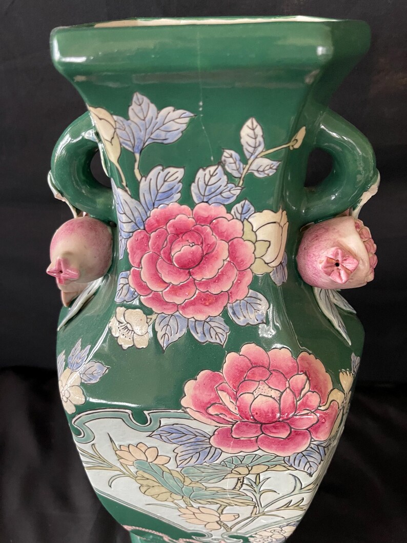 May include: A green ceramic vase with floral designs and two handles. The vase is decorated with pink and white flowers and green leaves. The handles are shaped like pomegranates.