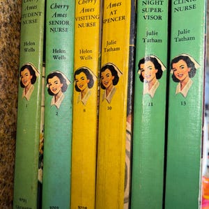 May include: A collection of vintage "Cherry Ames" novels. The books have colorful spines in shades of green and yellow, with illustrations of a nurse. Titles include "Student Nurse," "Senior Nurse," and "Clinic Nurse."